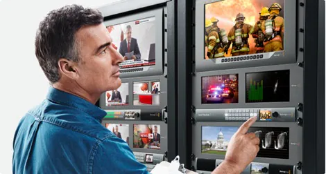 A man interacts with a control panel showing various video feeds including news and emergency scenes.
